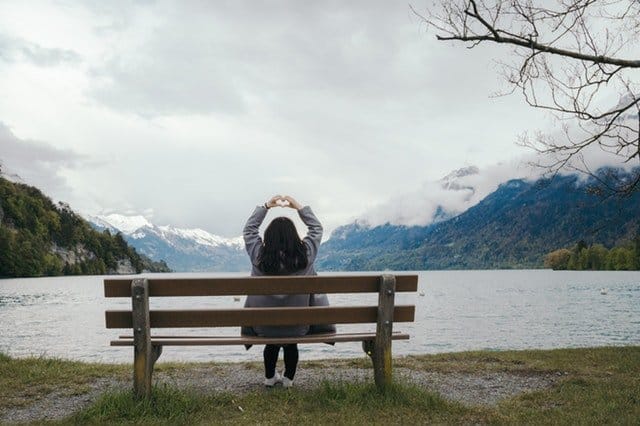 Girl on a bench with her back turned looking out at the mountains and making a heart with her hands