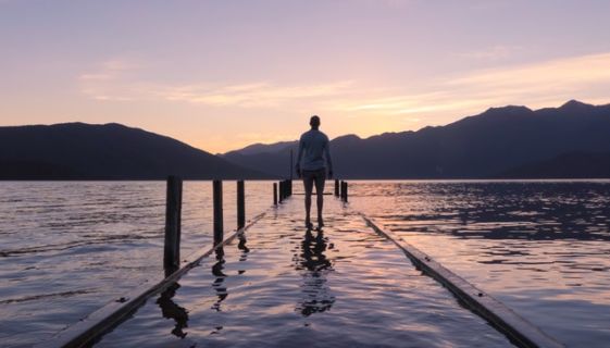 Person standing with their back turned on a flooded dock looking at the sunset and mountains